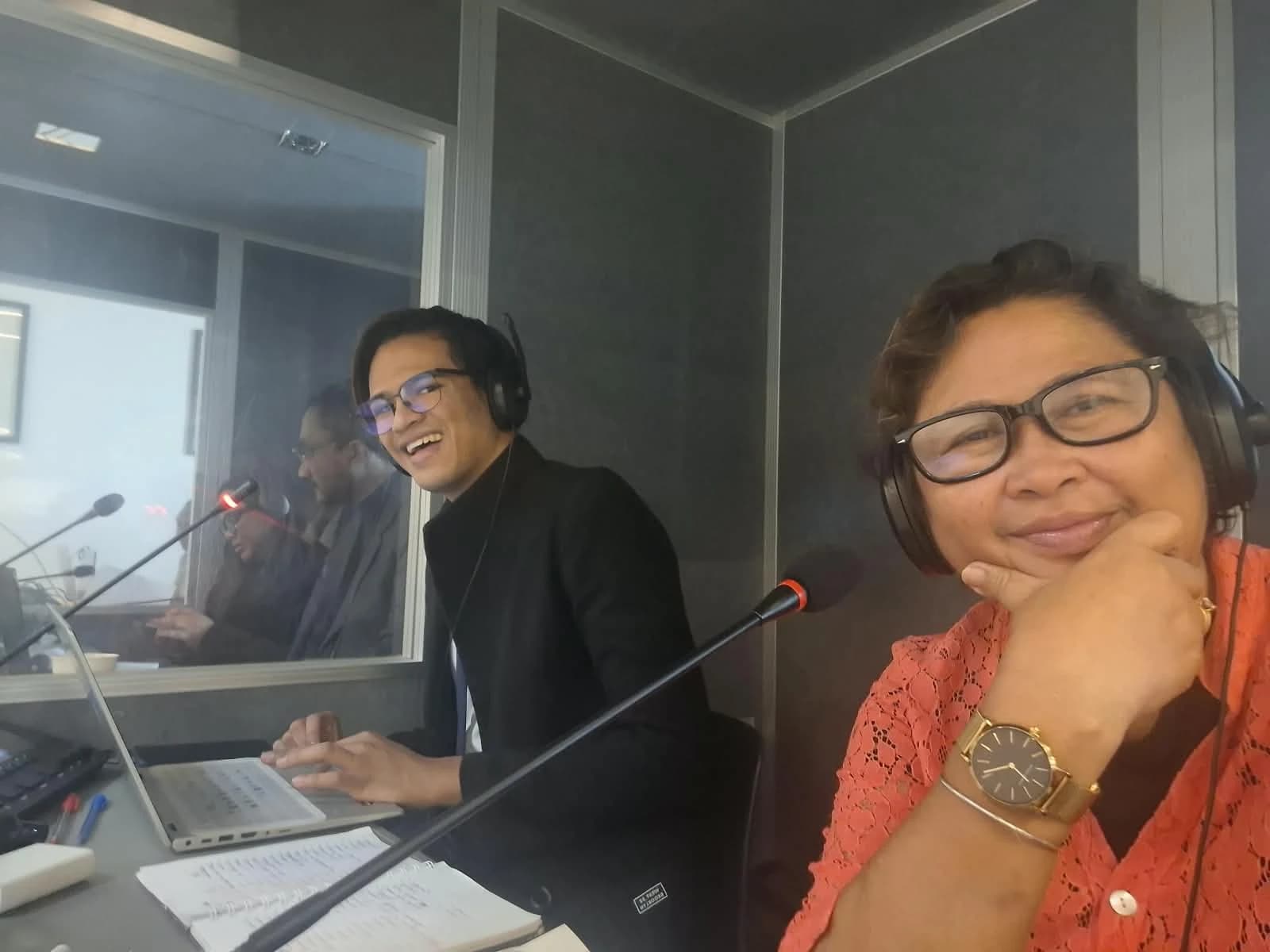 Two people wearing headsets beside microphones inside an interpretation booth